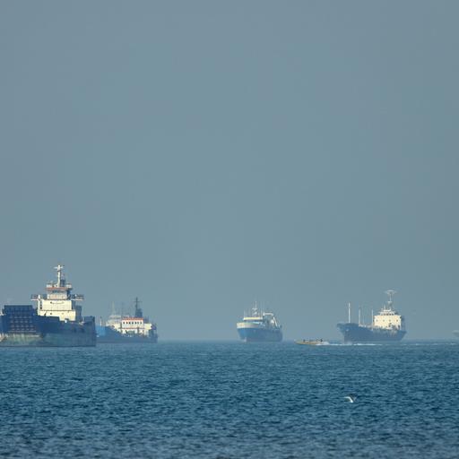 Frachtschiffe, darunter Massengutfrachter und Stückgutschiffe, liegen vor der Küste vor Anker, während im Vordergrund ein kleines Motorboot vorbeifährt 