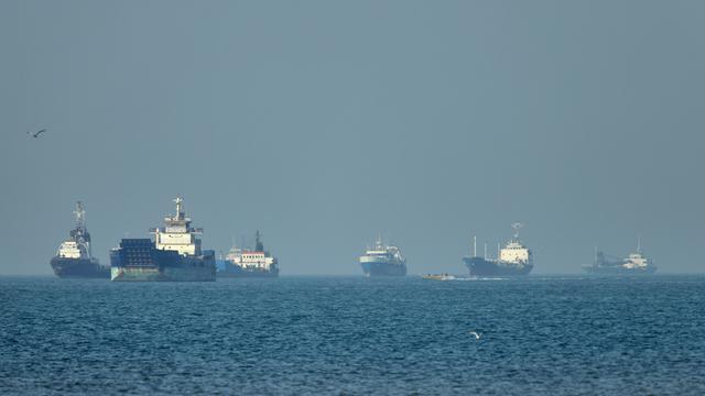 Frachtschiffe, darunter Massengutfrachter und Stückgutschiffe, liegen vor der Küste vor Anker, während im Vordergrund ein kleines Motorboot vorbeifährt 