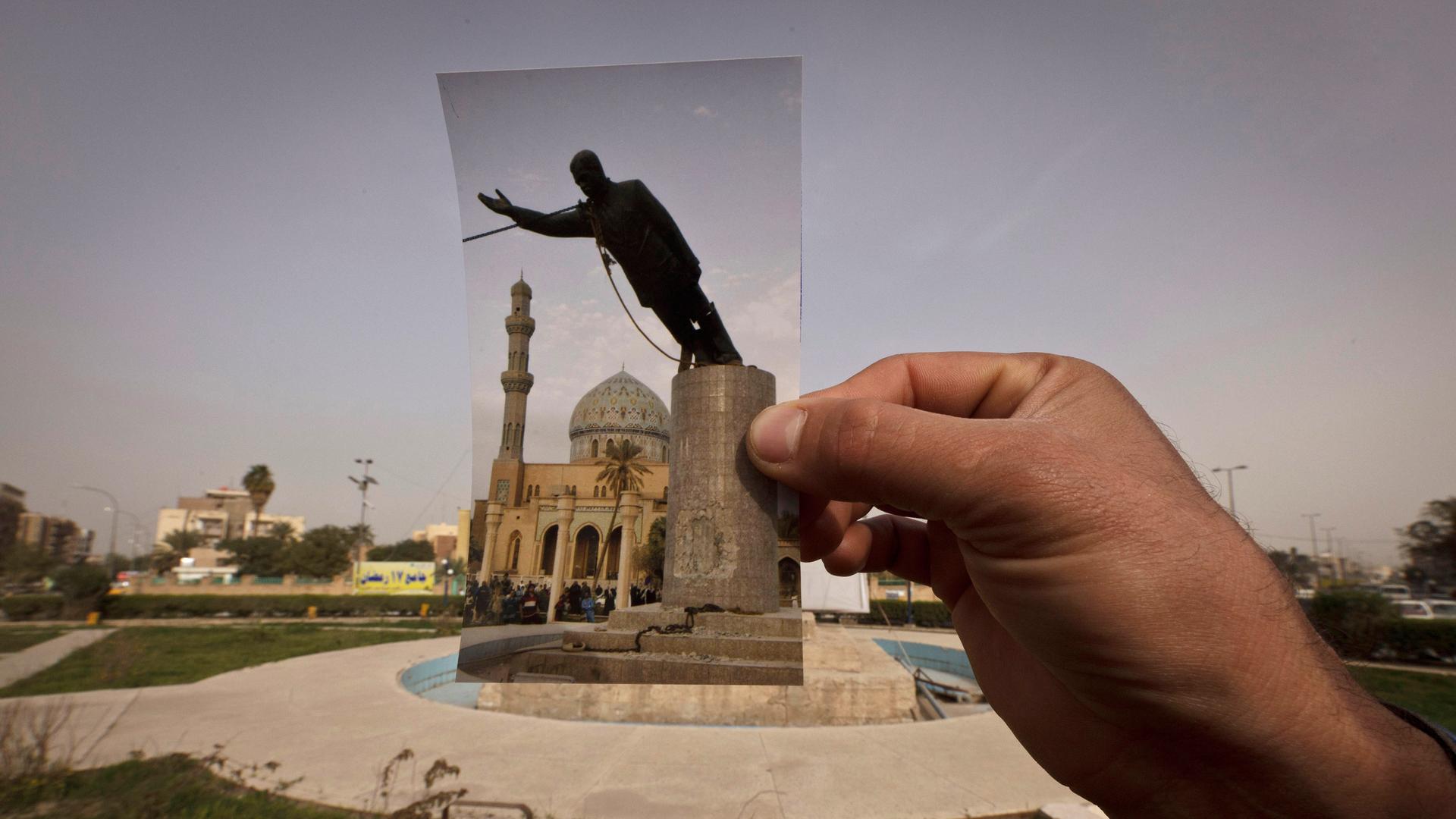 Eine Hand hält ein Foto an dem Platz, wo das Denkmal von Saddam Hussein 2003 gestürzt wurde.