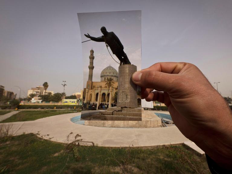 Eine Hand hält ein Foto an dem Platz, wo das Denkmal von Saddam Hussein 2003 gestürzt wurde.