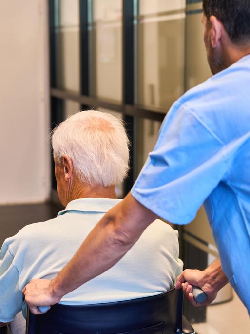 Ein Pfleger schiebt einen älteren Mann im Rollstuhl durch ein Gebäude. Ein Pfleger schiebt einen älteren Mann im Rollstuhl durch ein Gebäude.
