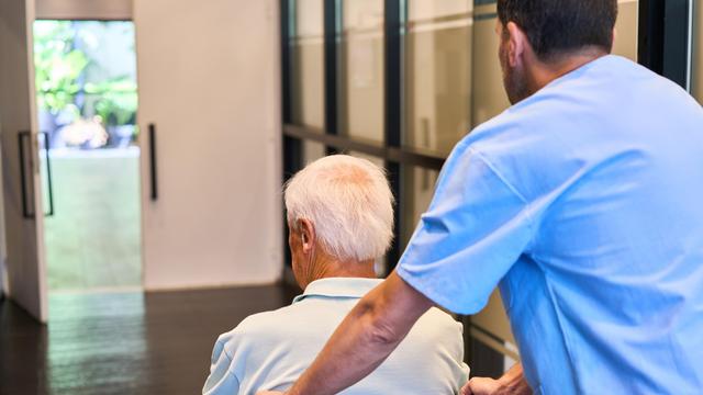 Ein Pfleger schiebt einen älteren Mann im Rollstuhl durch ein Gebäude.
