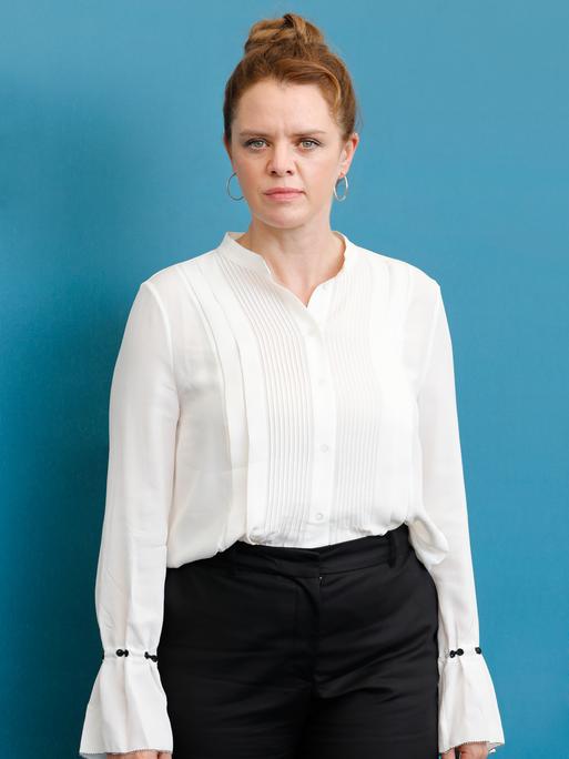 Julia von Heinz steht mit weißer Bluse und schwarzer Hose vor einem hellblauen Hintergrund und schaut in die Kamera