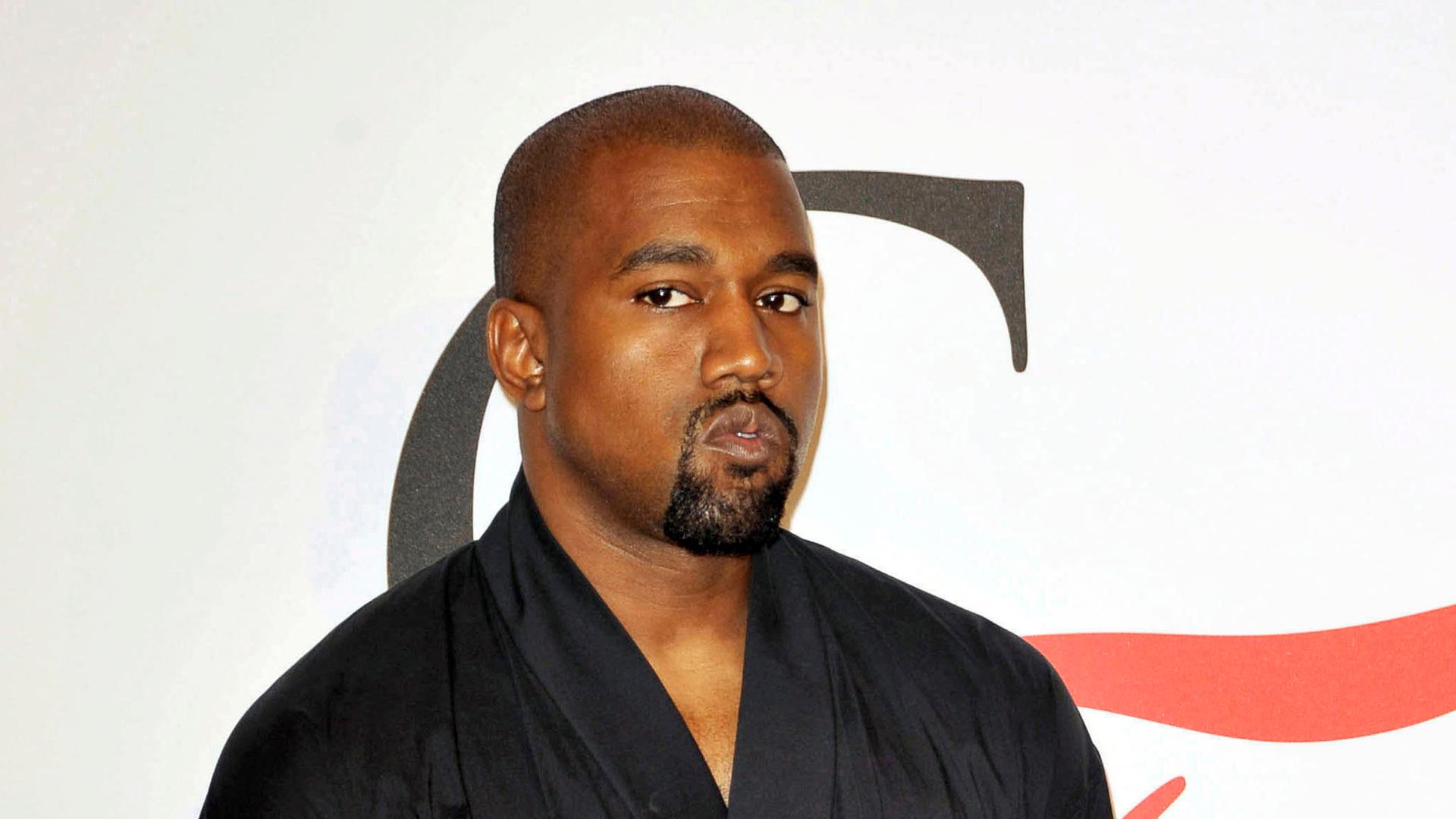 Der Rapper Kanye West schaut kritisch in die Kamera, er steht mit einem schwarzen Oberteil, das an einen Bademantel erinnert, vor einer weißen Wand.