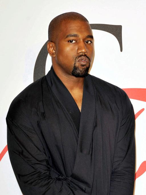 Der Rapper Kanye West schaut kritisch in die Kamera, er steht mit einem schwarzen Oberteil, das an einen Bademantel erinnert, von einer weißen Wand