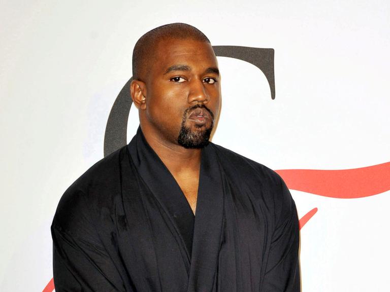 Der Rapper Kanye West schaut kritisch in die Kamera, er steht mit einem schwarzen Oberteil, das an einen Bademantel erinnert, von einer weißen Wand