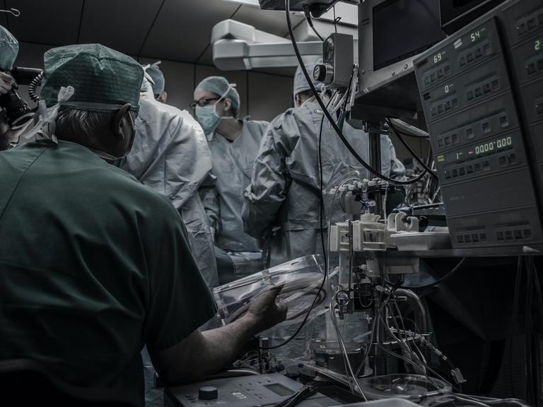 Ärzte in OP-Kitteln drängen sich im OP Saal, zu sehen ist außerdem eine Maschine mit vielen Kabeln.