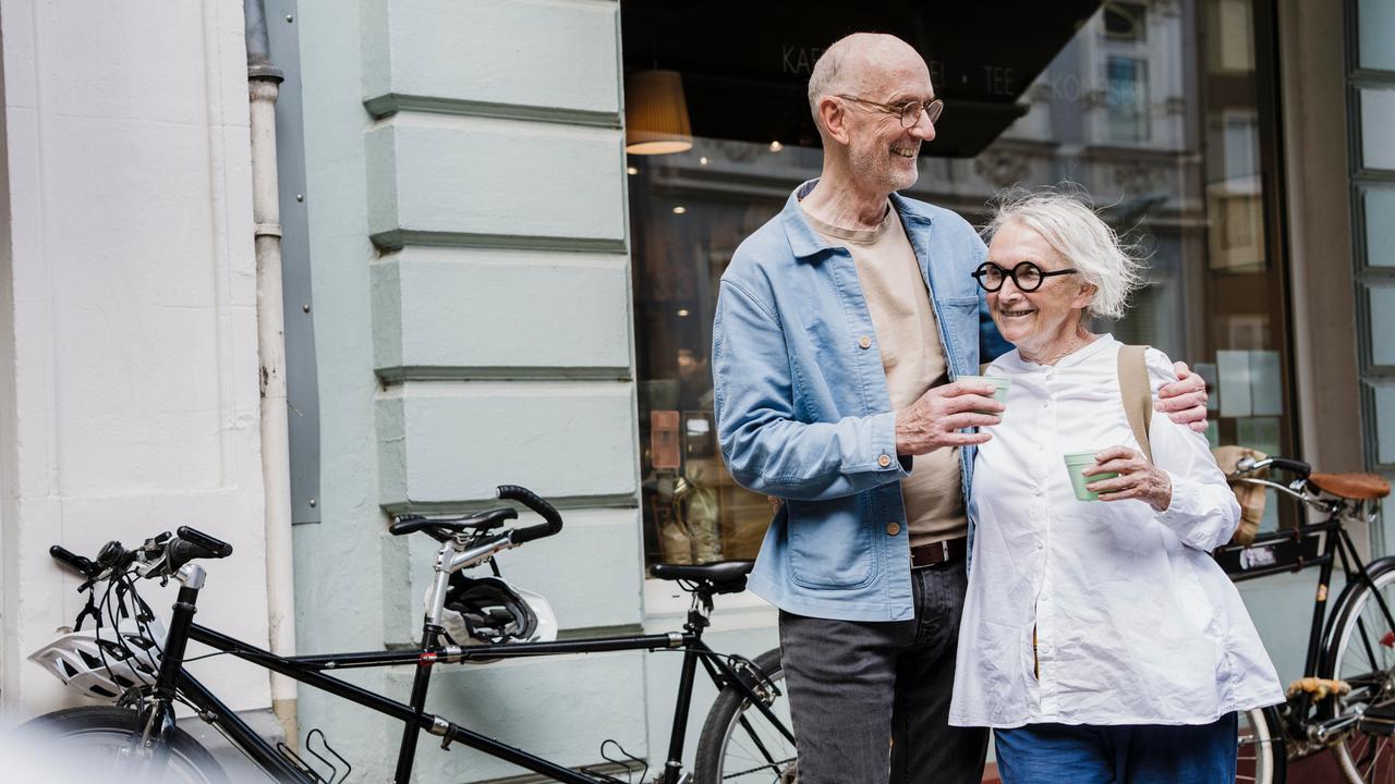 Ein glückliches Paar mit weißem Haar steht mit einem Kaffeebecher in der Hand vor einem Schaufenster, im Hintergrund stehen Fahrräder