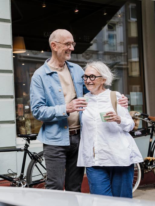 Ein glückliches Paar mit weißem Haar steht mit einem Kaffeebecher in der Hand vor einem Schaufenster, im Hintergrund stehen Fahrräder