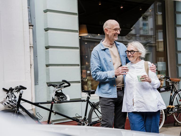 Ein glückliches Paar mit weißem Haar steht mit einem Kaffeebecher in der Hand vor einem Schaufenster, im Hintergrund stehen Fahrräder