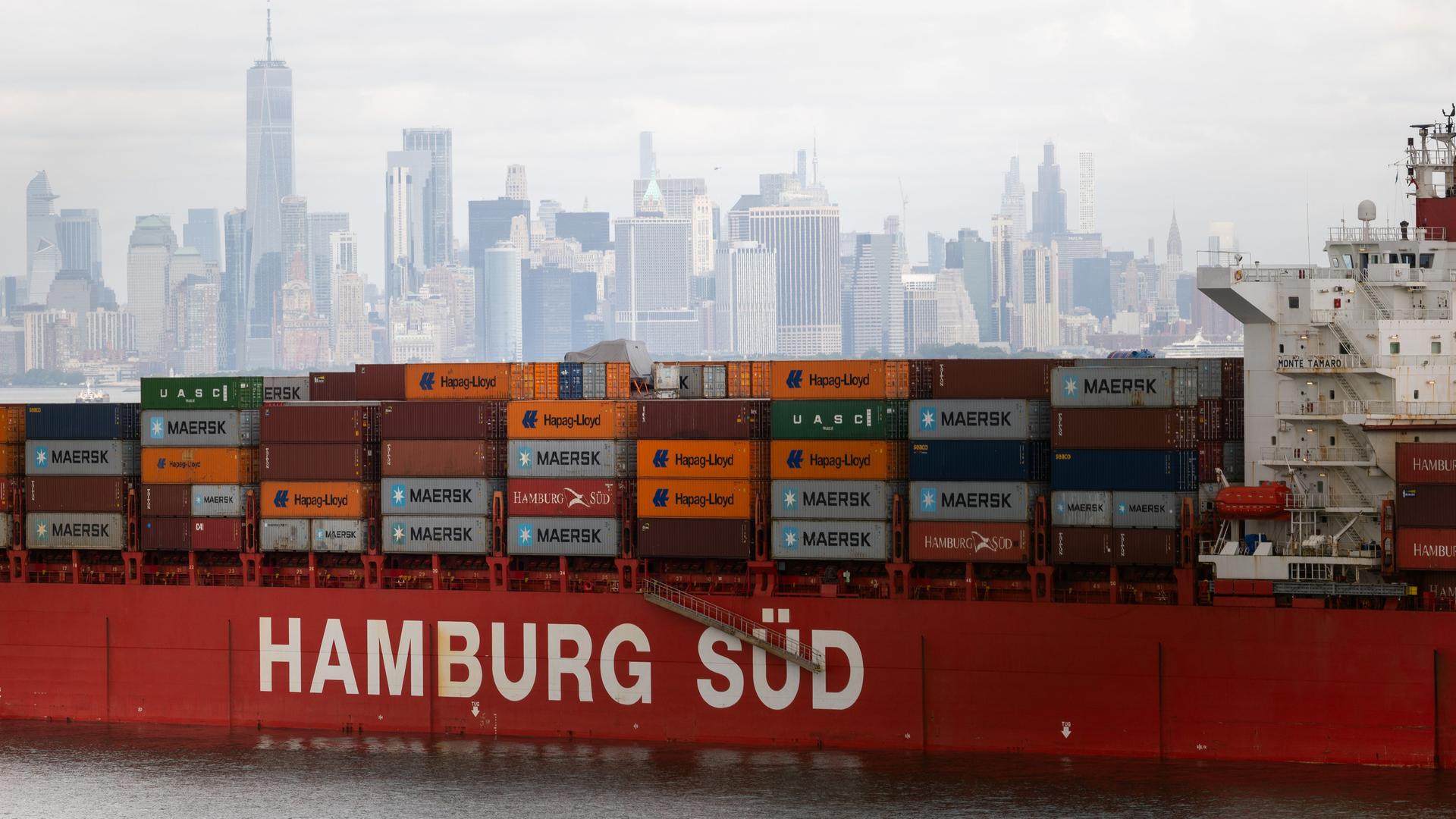 Ein Containerschiff liegt im New Yorker Hafen vor Anker. Es trägt die Aufschrift "Hamburg Süd". Im Hintergrund ist die Skyline von New York zu sehen.