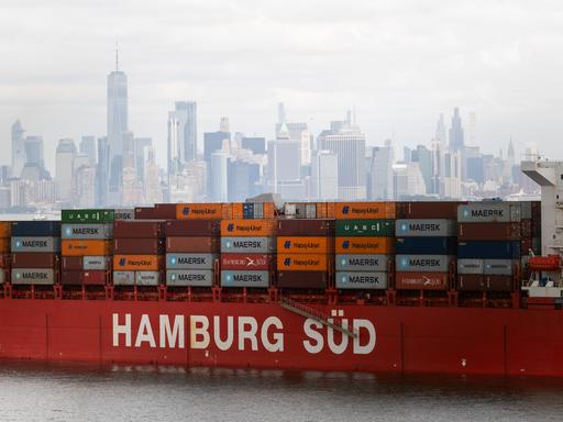 Ein Containerschiff liegt im New Yorker Hafen vor Anker. Es trägt die Aufschrift "Hamburg Süd". Im Hintergrund ist die Skyline von New York zu sehen.