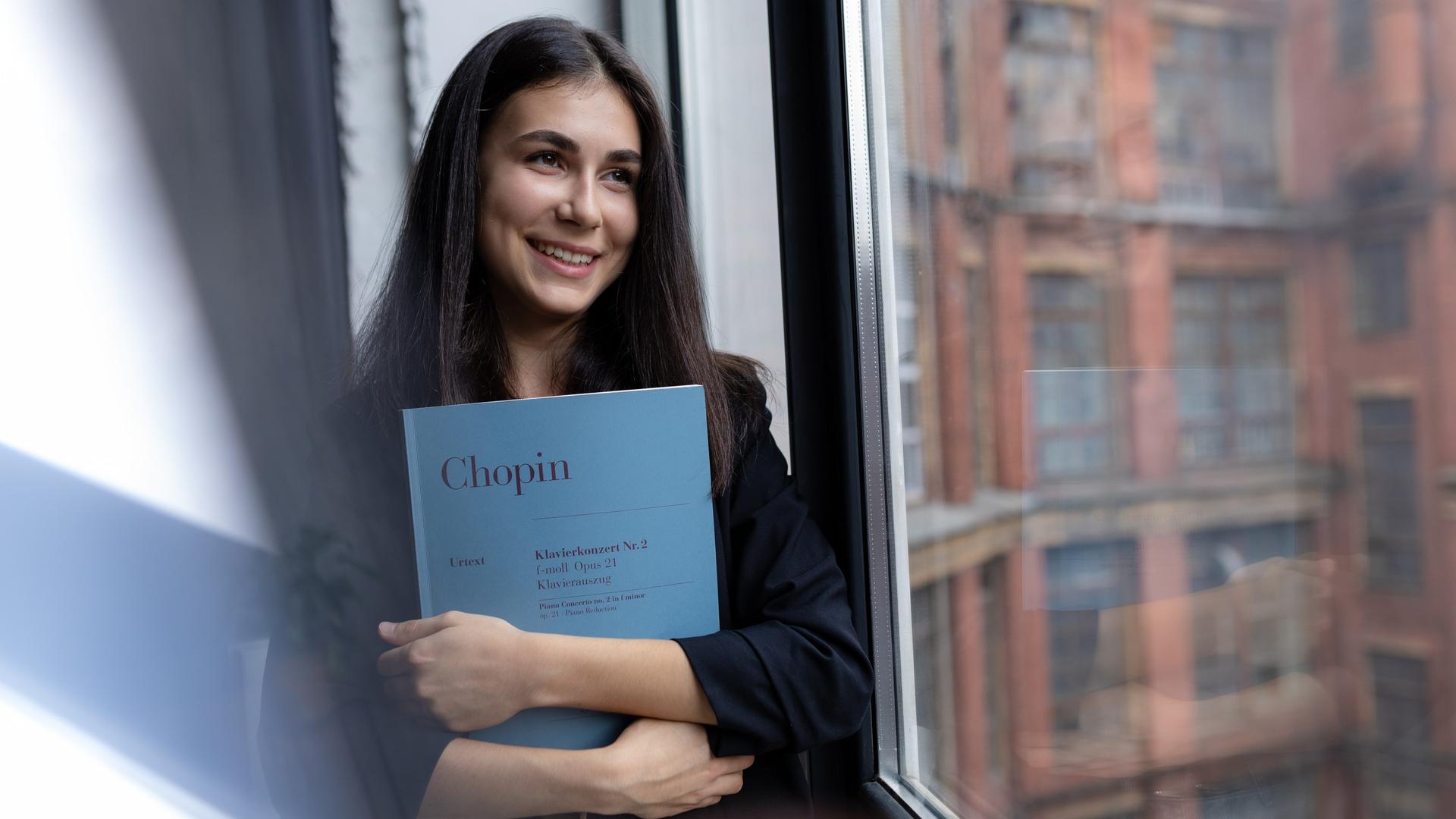 Eine junge Frau mit braunen Haaren hält die Partitur von Chopins Klavierkonzert Nr.2 im Arm und schaut lächelnd aus einem Fenster. Eine junge Frau mit braunen Haaren hält die Partitur von Chopins Klavierkonzert Nr.2 im Arm und schaut lächelnd aus einem Fenster.