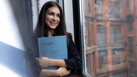 Eine junge Frau mit braunen Haaren hält die Partitur von Chopins Klavierkonzert Nr.2 im Arm und schaut lächelnd aus einem Fenster.