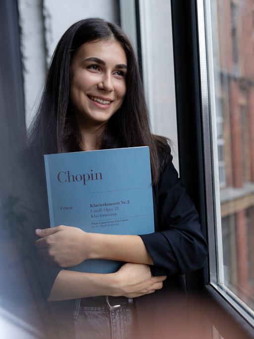 Eine junge Frau mit braunen Haaren hält die Partitur von Chopins Klavierkonzert Nr.2 im Arm und schaut lächelnd aus einem Fenster.