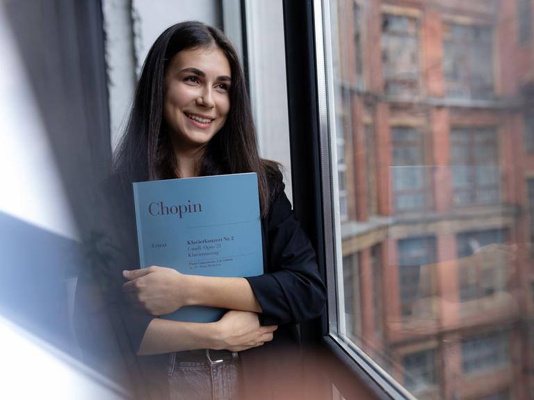 Eine junge Frau mit braunen Haaren hält die Partitur von Chopins Klavierkonzert Nr.2 im Arm und schaut lächelnd aus einem Fenster.