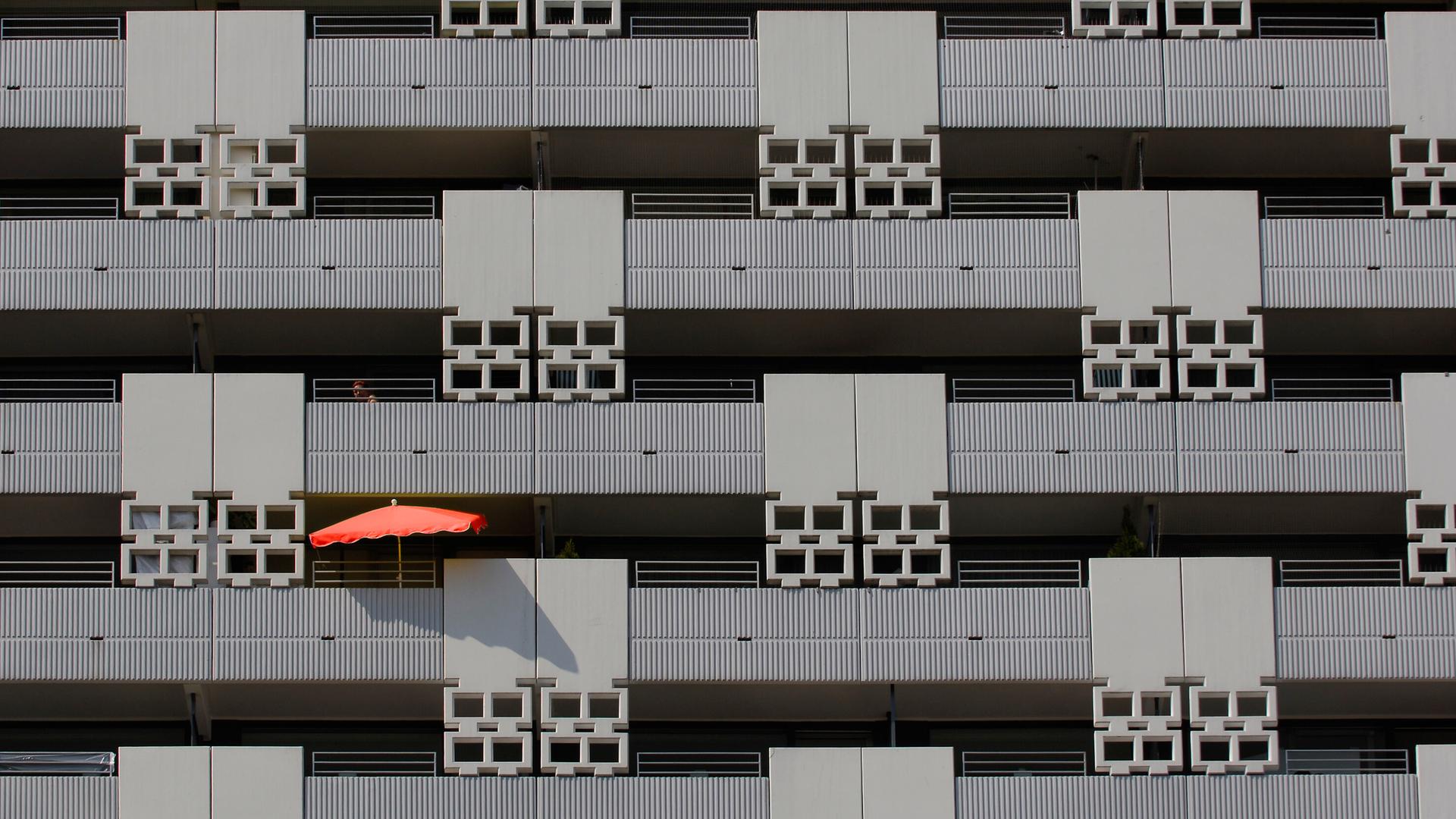 Blick auf eine grauen Häuser-Fassade mit mehreren Balkons - auf einem davon ist ein roter Sonnenschirm zu sehen.