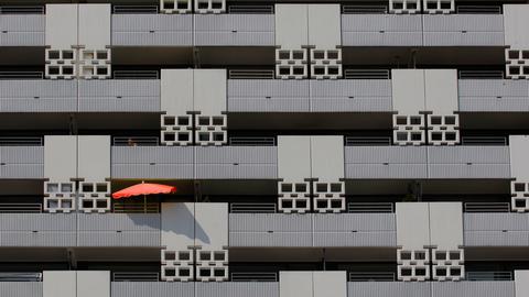 Blick auf eine grauen Häuser-Fassade mit mehreren Balkons - auf einem davon ist ein roter Sonnenschirm zu sehen.