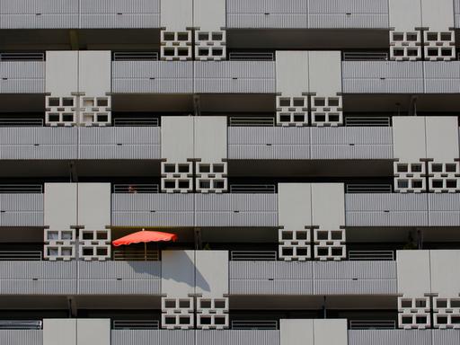 Blick auf eine grauen Häuser-Fassade mit mehreren Balkons - auf einem davon ist ein roter Sonnenschirm zu sehen.