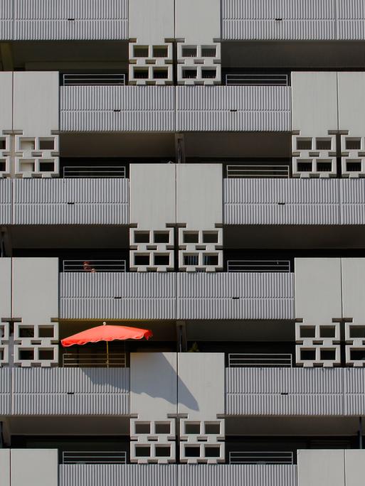 Blick auf eine grauen Häuser-Fassade mit mehreren Balkons - auf einem davon ist ein roter Sonnenschirm zu sehen.