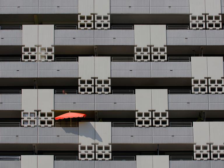 Blick auf eine grauen Häuser-Fassade mit mehreren Balkons - auf einem davon ist ein roter Sonnenschirm zu sehen.