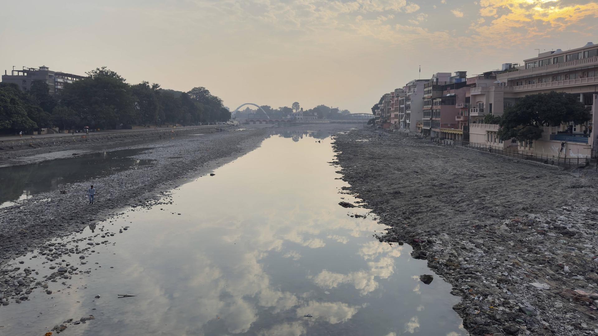 Flussbett des Ganges mit niedrigem Wasserstand und Spiegelungen im Wasser, im Hintergrund Gebäude und eine Brücke.