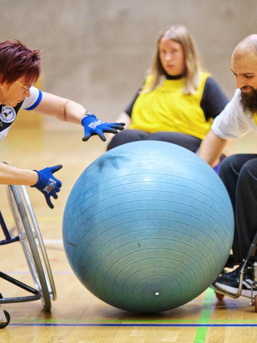 Menschen spielen Rollstuhl-Fußball. Sie schubsen mit den Händen einen sehr großen Ball.