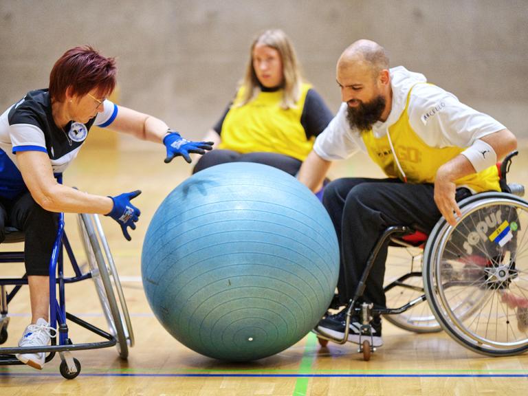 Menschen spielen Rollstuhl-Fußball. Sie schubsen mit den Händen einen sehr großen Ball.