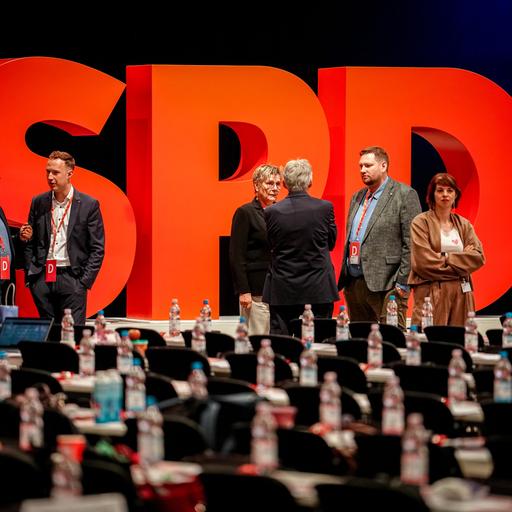Auf der Bühne vor einem SPD-Schriftzug aus stehenden großen roten Buchstaben stehen mehrere Teilnehmer und reden. Im Vordergrund unscharf die Delegiertenbänke. 