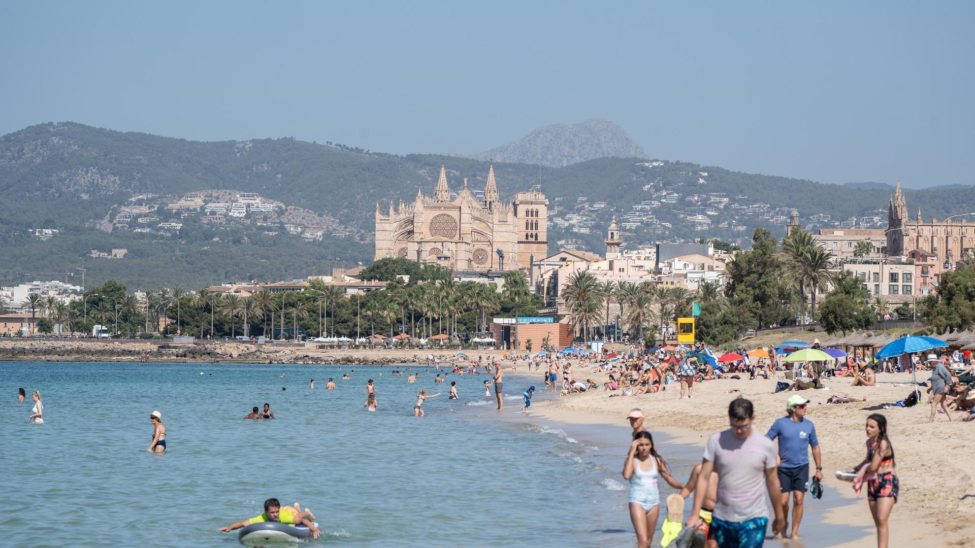 Die Mittelmeerinsel Mallorca im Juli 2024. Menschen sind am Strand; im Hintergund ist die Kathedrale zu sehen.