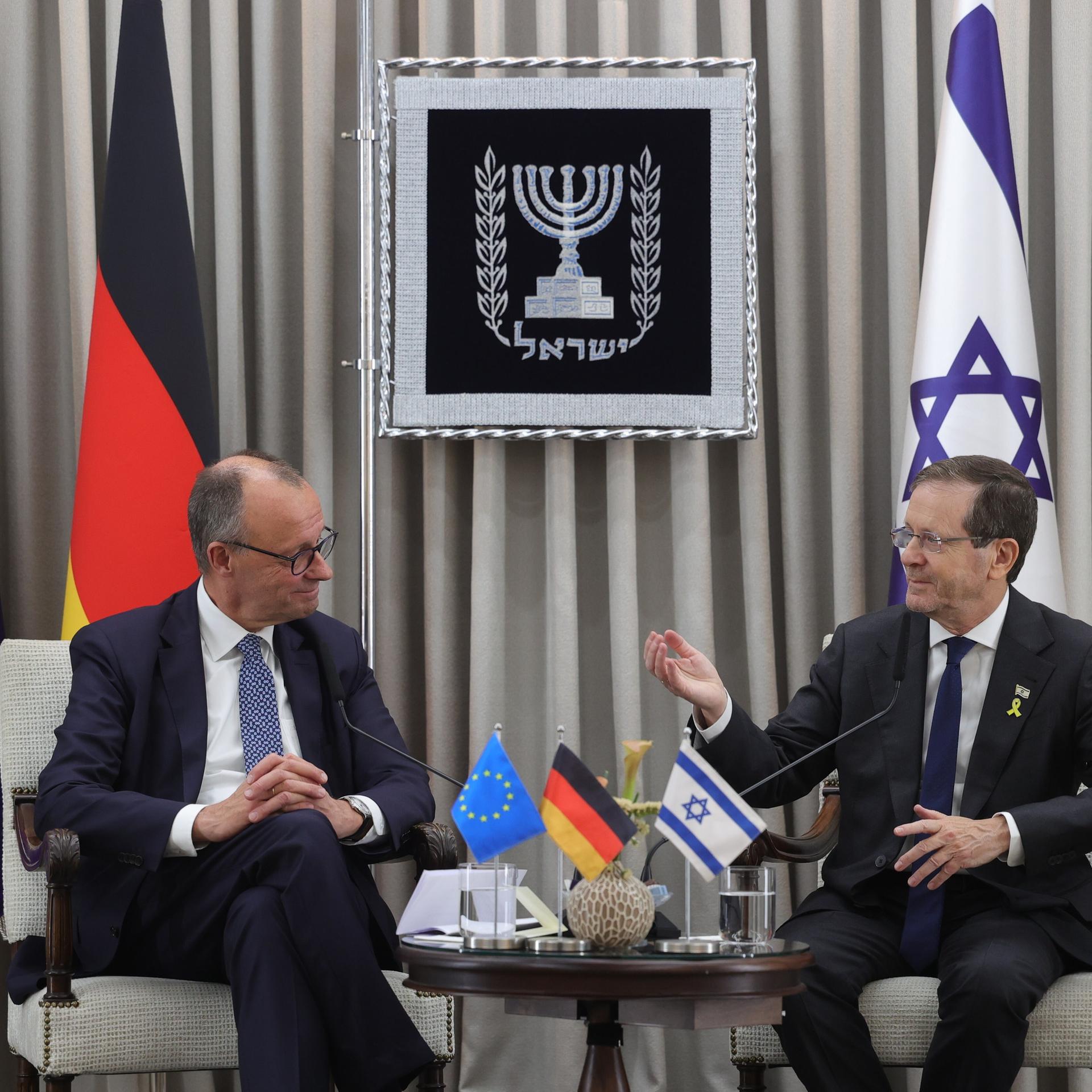 Bundeskanzler Merz und Israels Präsident Herzog sitzen nebeneinander und lachen. Hinter ihnen sind Fahnen von Deutschland, der EU und Israel zu sehen.