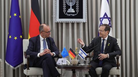 Bundeskanzler Merz und Israels Präsident Herzog sitzen nebeneinander und lachen. Hinter ihnen sind Fahnen von Deutschland, der EU und Israel zu sehen.