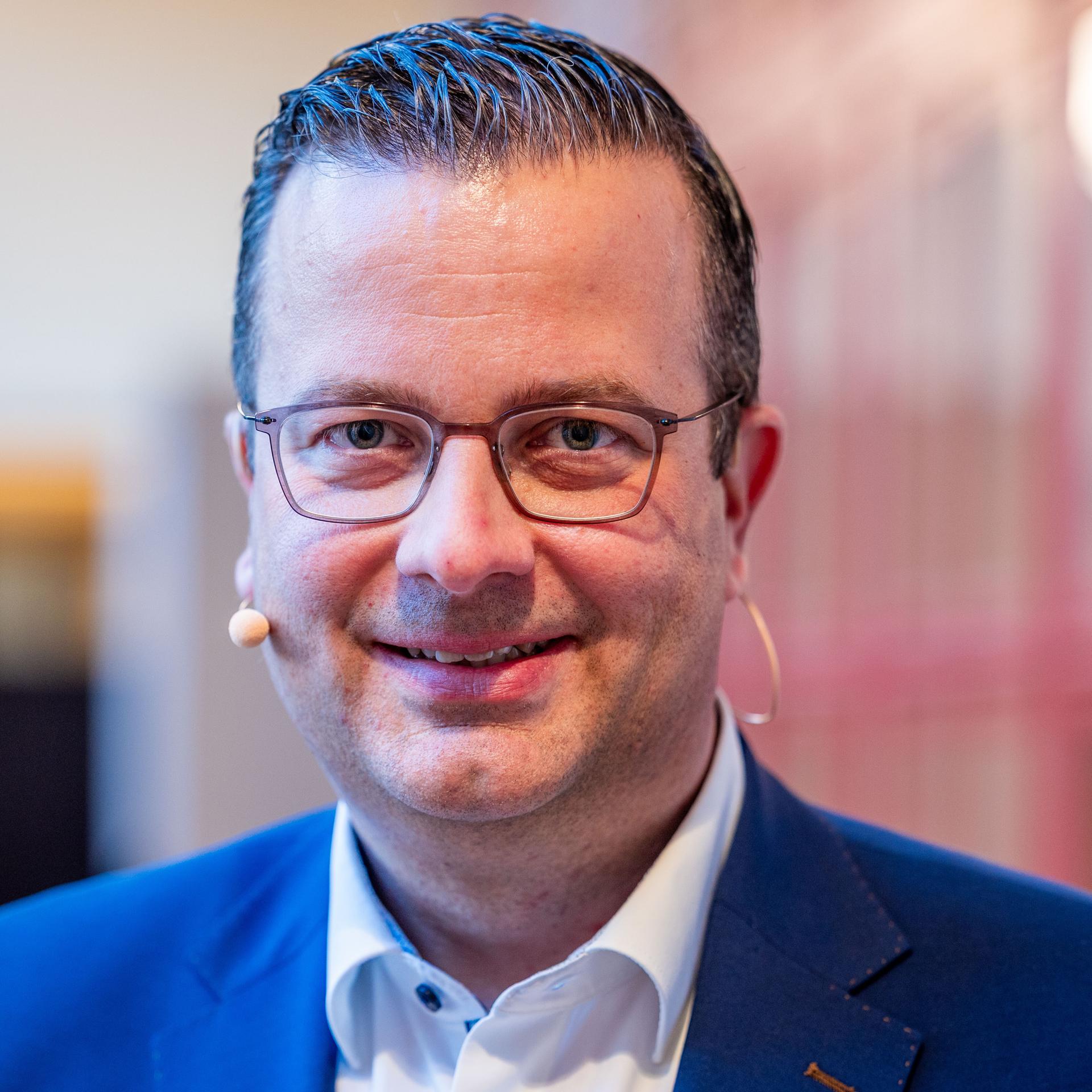 Ehlers blickt in die Kamera und lächelt. Er trägt eine Brille und ein Headset.