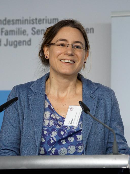 Katja Grieger steht an einem Pult und spricht gestikulierend und freundlich. Sie trägt ein blaues Sakko und eine blaugelb gemusterte Bluse.