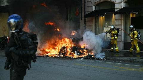 Am Straßenrand steht ein Fahrzeug in Flammen, Feuerwehrleute löschen den Brand. Ein Polizist mit Helm und Schutzkleidung steht in der Nähe.