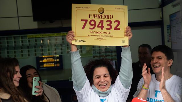 Eine Frau hält ein Schild hoch. Auf dem Schild steht eine Nummer. Mit dieser Nummer hat die Frau bei dem Lotto-Spiel viel Geld gewonnen. Die Frau freut sich darüber.