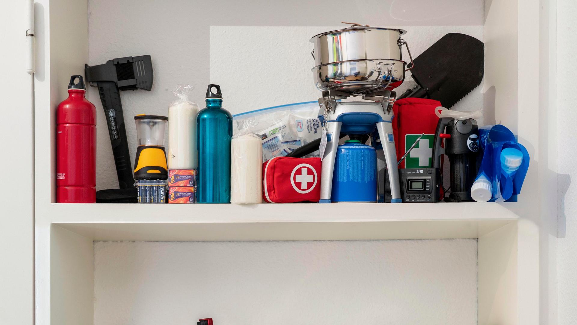 In einem Schrank eines privaten Haushalts sind verschiedene Vorräte gelagert, darunter Lebensmitteldosen, Kerzen, eine Axt, ein Gaskocher mit Kartusche, ein Notfallset sowie mehrere befüllbare Wasserbeutel.