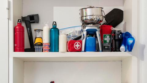 In einem Schrank eines privaten Haushalts sind verschiedene Vorräte gelagert, darunter Lebensmitteldosen, Kerzen, eine Axt, ein Gaskocher mit Kartusche, ein Notfallset sowie mehrere befüllbare Wasserbeutel.