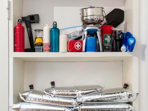 In einem Schrank eines privaten Haushalts sind verschiedene Vorräte gelagert, darunter Lebensmitteldosen, Kerzen, eine Axt, ein Gaskocher mit Kartusche, ein Notfallset sowie mehrere befüllbare Wasserbeutel.