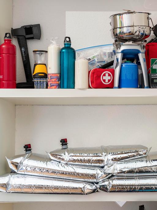 In einem Schrank eines privaten Haushalts sind verschiedene Vorräte gelagert, darunter Lebensmitteldosen, Kerzen, eine Axt, ein Gaskocher mit Kartusche, ein Notfallset sowie mehrere befüllbare Wasserbeutel.