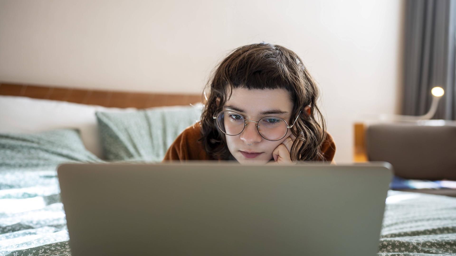 Eine junge Frau mit dunkelbraunen Haaren und markantem Pony, die entspannt in Bauchlage auf einem Bett liegt und konzentriert auf den Bildschirm ihres Laptops schaut.
