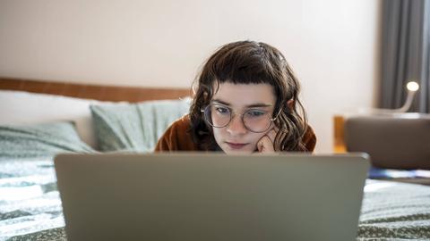 Eine junge Frau mit dunkelbraunen Haaren und markantem Pony, die entspannt in Bauchlage auf einem Bett liegt und konzentriert auf den Bildschirm ihres Laptops schaut.