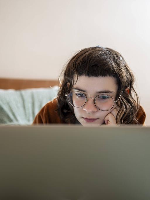 Eine junge Frau mit dunkelbraunen Haaren und markantem Pony, die entspannt in Bauchlage auf einem Bett liegt und konzentriert auf den Bildschirm ihres Laptops schaut. Eine junge Frau mit dunkelbraunen Haaren und markantem Pony, die entspannt in Bauchlage auf einem Bett liegt und konzentriert auf den Bildschirm ihres Laptops schaut.
