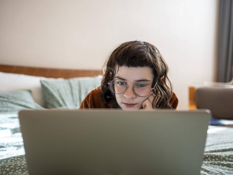 Eine junge Frau mit dunkelbraunen Haaren und markantem Pony, die entspannt in Bauchlage auf einem Bett liegt und konzentriert auf den Bildschirm ihres Laptops schaut. Eine junge Frau mit dunkelbraunen Haaren und markantem Pony, die entspannt in Bauchlage auf einem Bett liegt und konzentriert auf den Bildschirm ihres Laptops schaut.