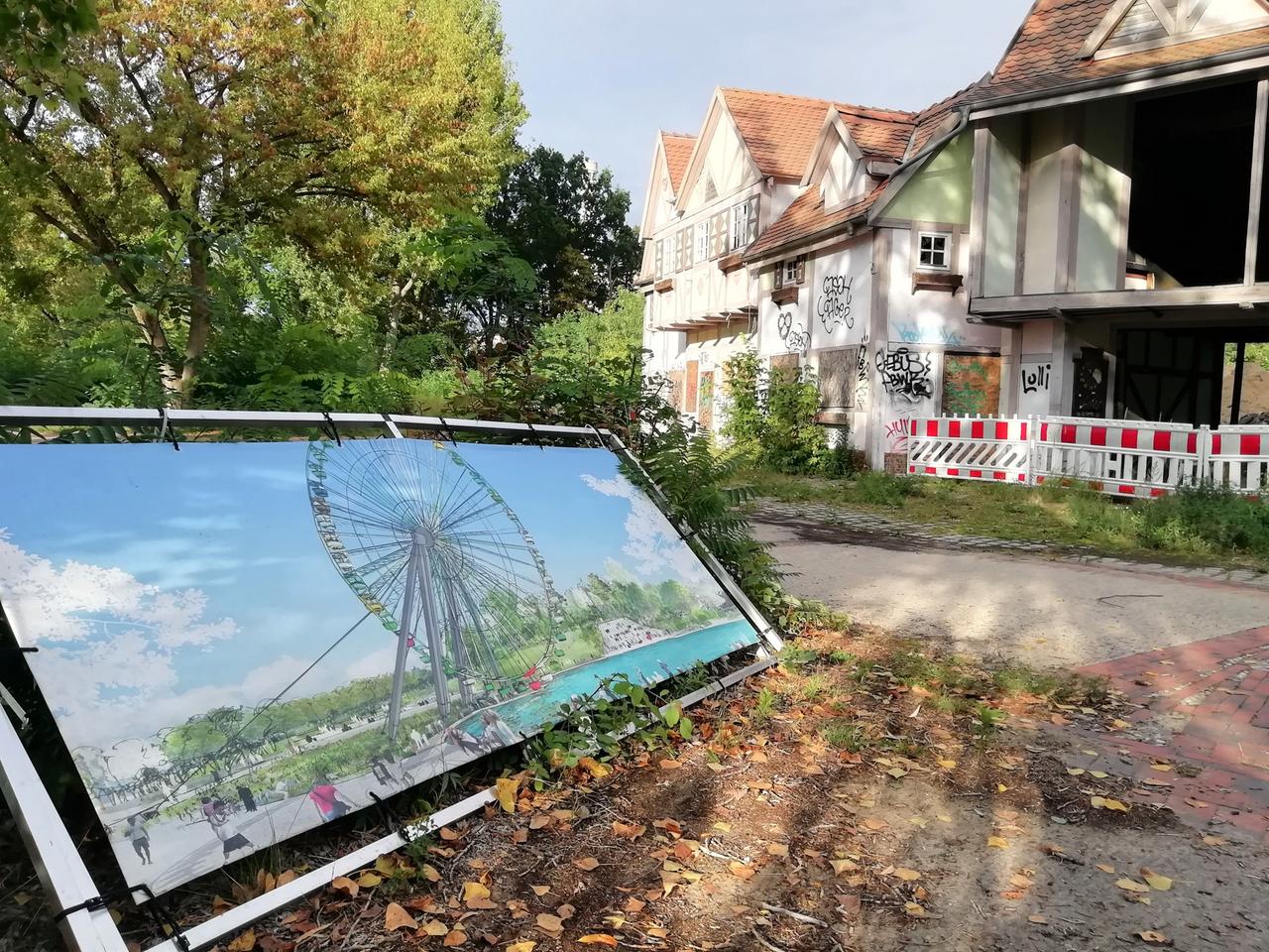 Auf dem Gelände des Spreeparks ist eine Illustration aufgestellt im Querformat in einem Rahmen gespannt und aufgestellt. Im Hintergrund sind Bäume und Häuser auf dem Gelände zu sehen, bei einem Haus fehlen ganze Wände.