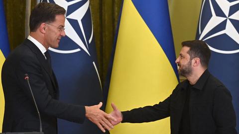 NATO-Genralsekretär Rutte und der ukrainische Präsident Selenskyj reichen sich die Hand. Hinter ihnen die NATO- und die Ukraine-Flagge.