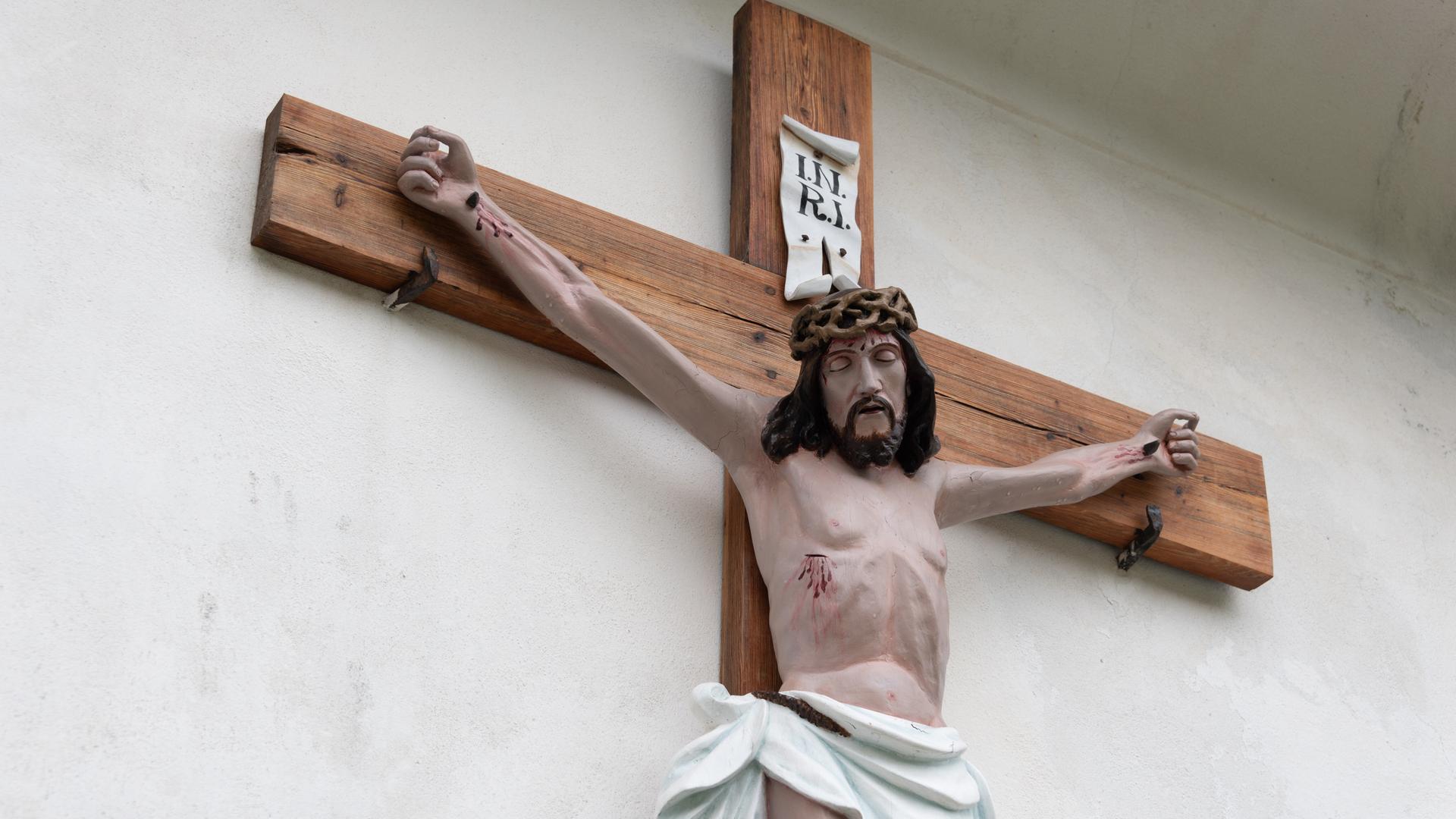 Ein Kruzifix hängt einer Außenwand einer Pfarrkirche, es zeigt eine farbig gefasste Darstellung des gekreuzigten Christus
