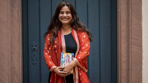 Porträt von Mithu Sanyal, die eine rot-orangene Jacke trägt und mit einem Buch in der Hand lachend vor einer schwarzen Tür steht.