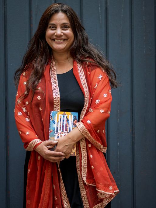 Porträt von Mithu Sanyal, die eine rot-orangene Jacke trägt und mit einem Buch in der Hand lachend vor einer schwarzen Tür steht.