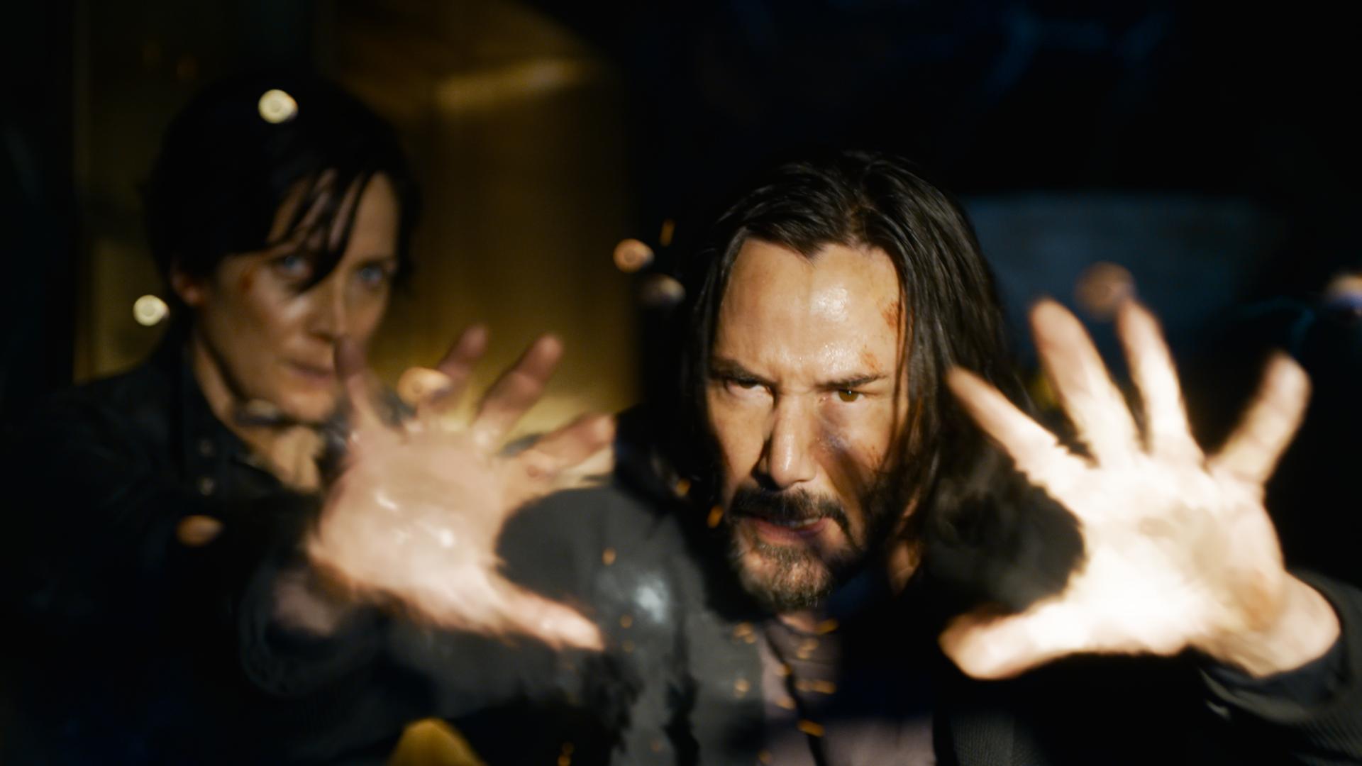 Keanu Reeves (Neo) und Carrie-Ann Moss (Trinity) in einer Szene des Films "Matrix Resurrections". Neo streckt die Hände einem unbekannten Gegner entgegen, Trinity blickt im Hintergrund furchtsam.